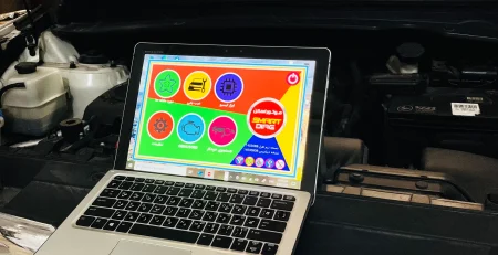 An open Kia vehicle hood with a laptop placed on the front grille. The laptop screen shows a colorful interface with various icons and Arabic text, likely related to vehicle diagnostics or maintenance