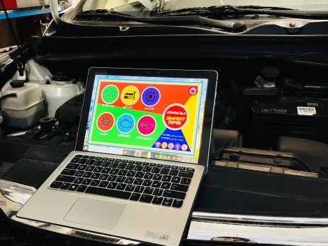 An open Kia vehicle hood with a laptop placed on the front grille. The laptop screen shows a colorful interface with various icons and Arabic text, likely related to vehicle diagnostics or maintenance