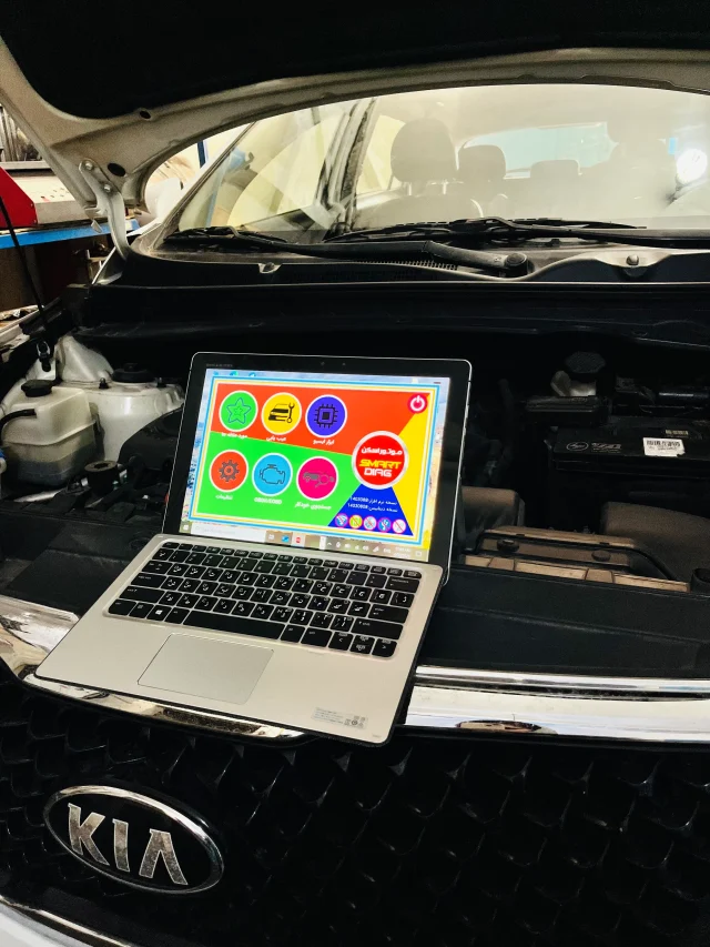 An open Kia vehicle hood with a laptop placed on the front grille. The laptop screen shows a colorful interface with various icons and Arabic text, likely related to vehicle diagnostics or maintenance